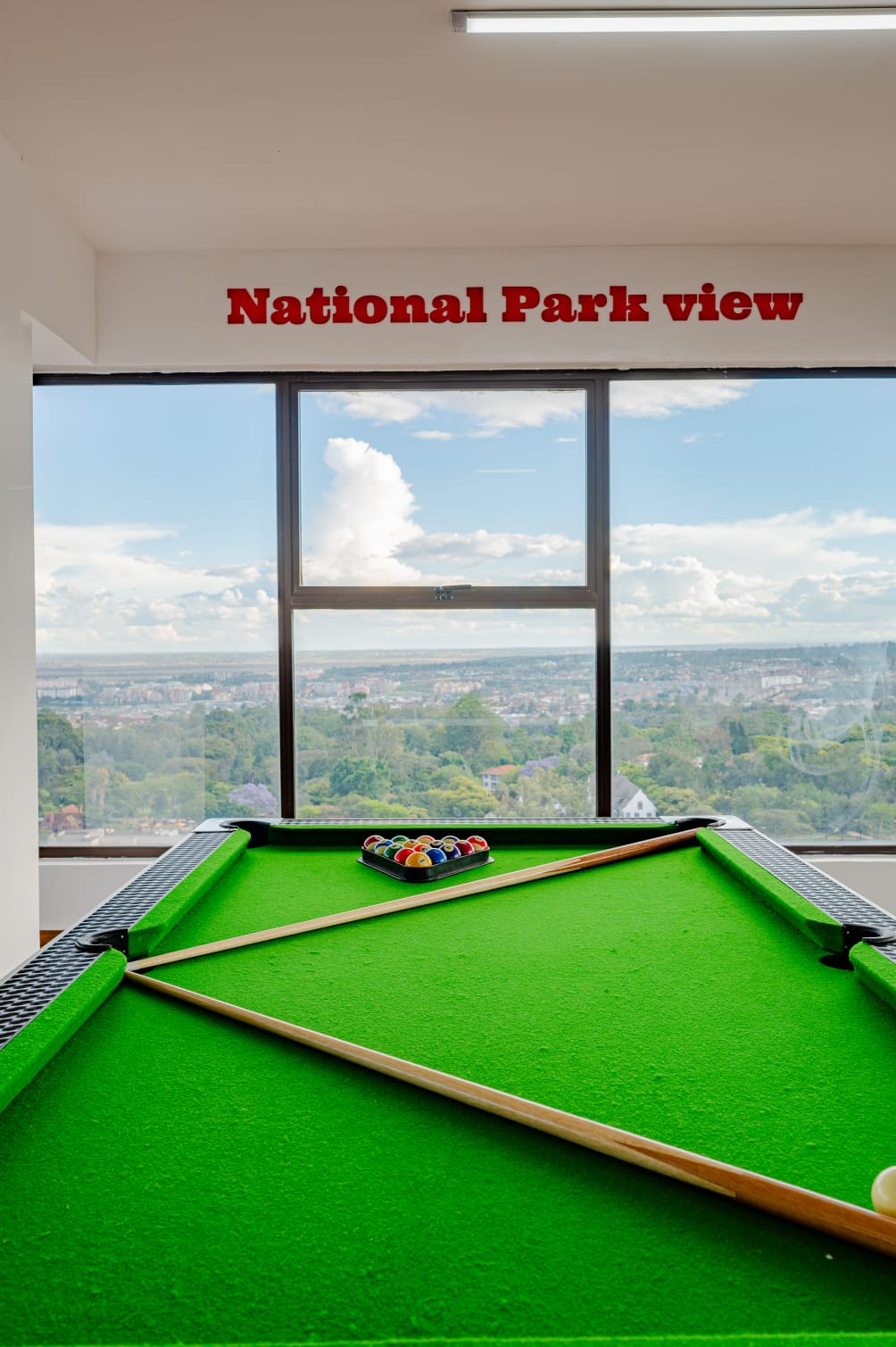 Green pool table with cues and racked balls beside large windows overlooking trees and buildings in Kilimani.