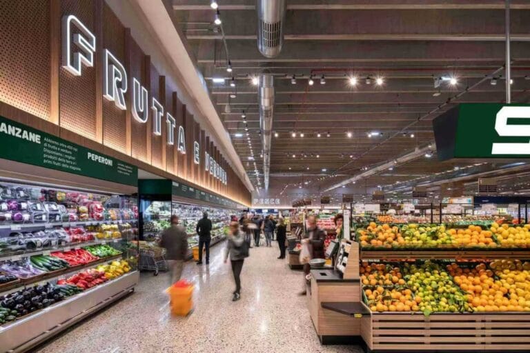 Supermarket produce aisle with shoppers, refrigerated vegetables and tiered displays of citrus fruit.